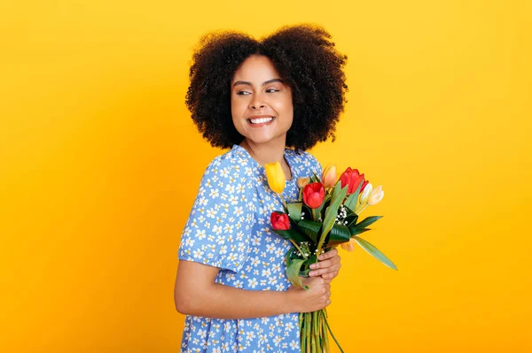 Lovely fashionable brazilian or african american woman, in a blue summer dress, holding bouquet of fresh multicolored tulips for a birthday or womens day, looks aside, isolated orange background