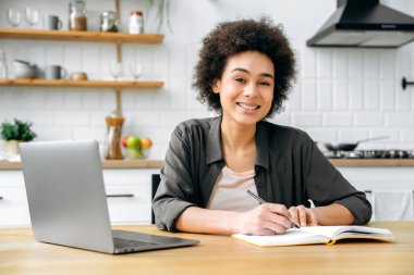 Güzel, mutlu, kıvırcık saçlı, öğrenci ya da serbest çalışan Afro-Amerikan bir kadın, bir dizüstü bilgisayarla mutfakta oturuyor, çalışıyor ya da çalışıyor, notlar alıyor, kameraya bakıyor, gülümsüyor.