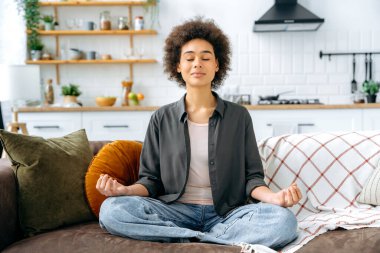 Rahatlama ve meditasyon konsepti. Rahat ve huzurlu bir Afro-Amerikan kadını. Günlük giysiler içinde. Oturma odasında kanepede tek başına oturuyor ve lotus pozisyonunda meditasyon yapıyor.