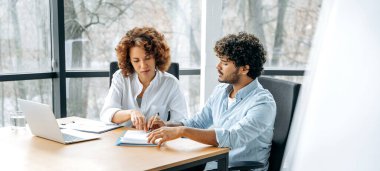 İki meslektaşın panoramik fotoğrafı, Afrikalı Amerikalı bir kadın ve Hintli bir adam, ofiste bir masada oturmuş, mali belgeleri inceliyor, yeni bir projenin risklerini ve beklentilerini analiz ediyorlar.