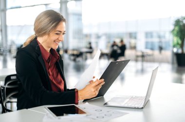 Modern bir ofiste masasında oturan, belgelerle ve dizüstü bilgisayarla bir projede çalışan, raporları, finansal grafikleri analiz eden, gülümseyen mutlu, kendinden emin, başarılı beyaz bir iş kadını.