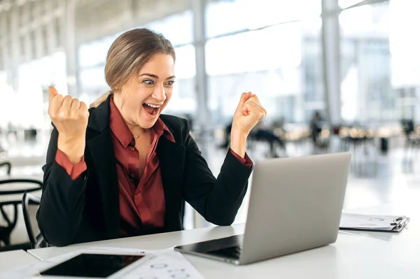 Joyful excited caucasian woman, manager, office employee, sits at a work desk in office, looks in amazement at the laptop screen, rejoices at success, good result, news, gesticulates with her hands