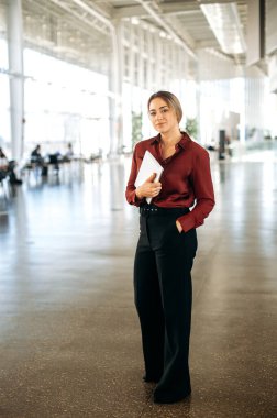 Kendine güveni tam, beyaz, takım elbiseli başarılı bir iş kadınının uzun bir fotoğrafı. Bir iş merkezinde elinde tabletle duruyor. Kameraya bakıyor ve gülümsüyor.