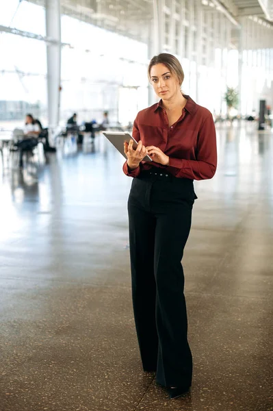 Full length photo of a positive elegant beautiful confident successful caucasian business lady dressed in a suit, stands in a coworking center with a tablet in her hands, looks at the camera