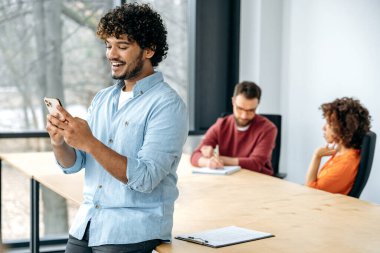 Neşeli, yakışıklı Hintli ya da Arap bir adam, ofis çalışanı, ofisteki bir masada oturuyor, akıllı telefonunu kullanıyor, mesajlaşıyor, iyi haberler okuyor, gülümsüyor, meslektaşları arka planda oturuyor.