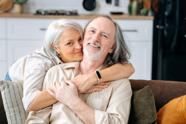 Orta yaşlı mutlu bir beyaz çiftin portresi, olgun eşler evdeki bir oturma odasında birlikte vakit geçiriyorlar, bir kadın kanepeye uzanıyor, kameraya bakıyor, gülümsüyor.