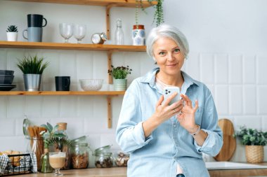Pozitif stil sahibi, olgun, beyaz saçlı bir kadın, mutfakta duruyor, akıllı telefonunu elinde tutuyor, sosyal ağlarda iletişim kurmak için kullanıyor, haberleri okuyor, kameraya bakıyor, gülümsüyor.