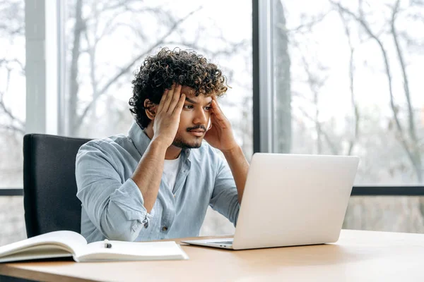 Sad worried indian or arabian curly man, financial manager, company ceo, sitting at a desk in the office, looking at a laptop screen in confusion, has work problems, experiencing anxiety, stress