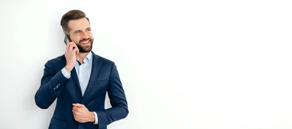 stock image Mobile phone conversation. Panoramic photo of caucasian business man in elegant suit, stands on a white isolated background, talking on the smartphone with a colleague, looking away, smile. Copy-space