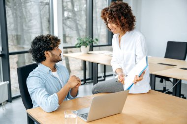 Farklı etnik kökenden iki pozitif iş ortağı, bir erkek ve bir kadın, ofiste birlikte çalışıyorlar, beyin fırtınası yapıyorlar, fikirler ve stratejiler tartışıyorlar, gülümsüyorlar. İşbirliği, ortaklık kavramı