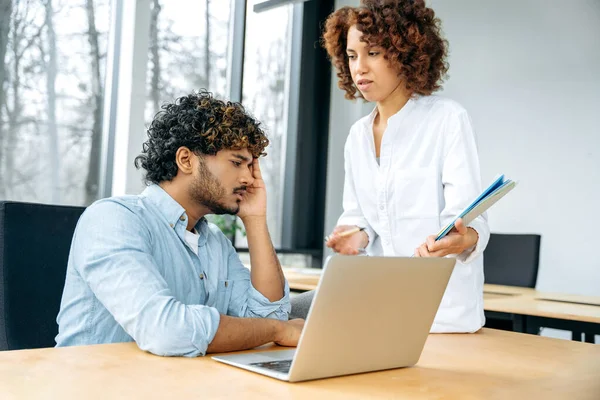 Indian man and african american woman, company colleagues, work together in the modern office, a woman came to a colleagues time to share ideas and suggestions, a man looks tired and worried