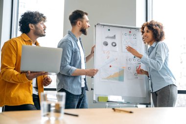 Çok ırklı finansal analistlerden oluşan bir ekip, ekonomi uzmanları, beyaz tahtaya yakın modern ofiste duruyor, finansal stratejiyi, planı, perspektifi, fikirleri ve düşünceleri paylaşıyor, gülümsüyor.
