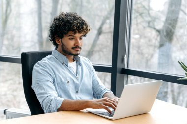 Zeki, kendine güvenen Hintli veya Arap bir adam, şirket seosu, bilişim uzmanı, İK yöneticisi, ofiste çalışma masasında otururken dizüstü bilgisayara bakıyor, gülümsüyor.