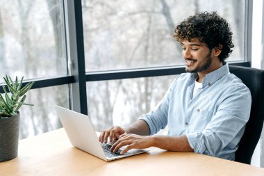 Pozitif Hintli ya da Arap iş adamı, şirket çalışanı, CEO, yönetici, ofiste oturuyor, bir dizüstü bilgisayar üzerinde çalışıyor, yeni bir proje planlıyor, strateji geliştiriyor ve tanıtımını yapıyor.