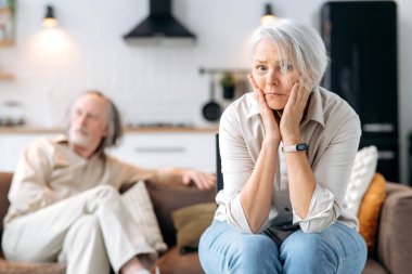 Senior caucasian spouses misunderstanding each other, experiencing crisis in relationship, offended wife looks disappointedly at camera, husband sits in defocus on background. Quarrel, marital crisis clipart