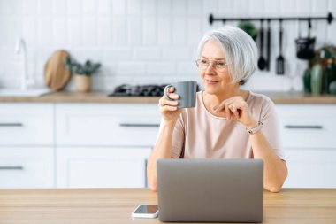 Güzel, pozitif, beyaz, gözlüklü bir kadın, evinde dizüstü bilgisayarla bir masada oturuyor, internette çalışıyor, kahve molası veriyor, dinleniyor, uzaklara bakıyor, düşünüyor, hayal kuruyor, gülümsüyor.
