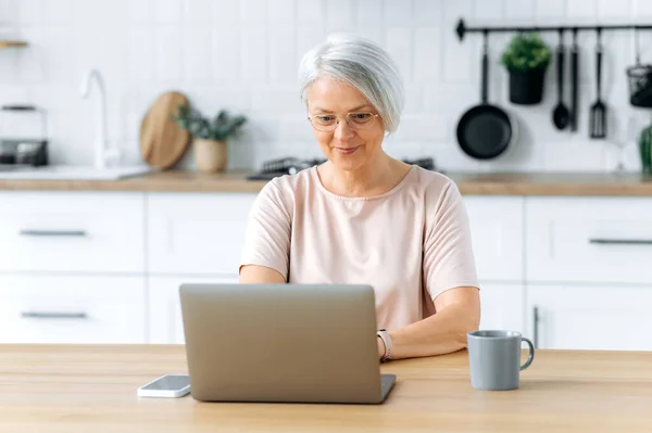 stock image Positive pretty modern caucasian middle aged gray haired woman with glasses, sits at home in the kitchen, uses laptop for remote work, online shopping, read news, looks satisfied, smiles