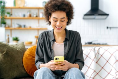 Cihazları kullanarak. Sevimli, Afro-Amerikan, gündelik giysiler içinde, arka planda bir kanepeye oturmuş, akıllı telefon kullanıyor, sosyal medyada sohbet ediyor, mesajlaşıyor, gülümsüyor.