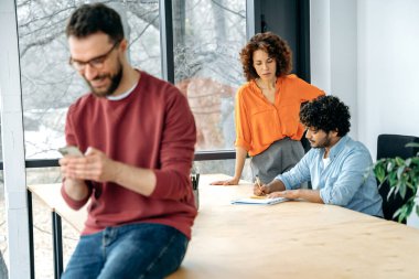 Fosforlu modern beyaz erkek, yaratıcı yönetici, seo, ofisteki bir masada otururken akıllı telefon kullanıyor, internette geziniyor, mesajları cevaplıyor, iş arkadaşları arka planda oturuyor.