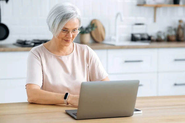 stock image Modern positive gray haired middle aged caucasian woman with glasses, sitting at a table at home in the kitchen with a laptop, working remotely, texting with a client, working on a project, smile