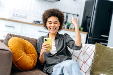 Başarı, zafer konsepti. Neşeli, modaya uygun, Afro-Amerikan genç kadın, oturma odasında bir kanepede oturuyor, akıllı telefon kullanıyor, el kol hareketi yapıyor, başarıyla seviniyor, zafer, harika haberler var, mutlu bir şekilde gülümsüyor.