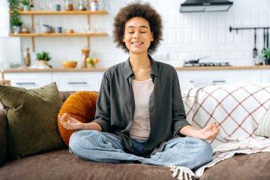 Sakin, huzurlu, rahat, rahat, rahat, rahat, gündelik giysiler içinde, evinde, kanepede tek başına oturmuş, lotus pozisyonunda meditasyon yapıyor, gülümsüyor. Rahatlama ve meditasyon kavramı
