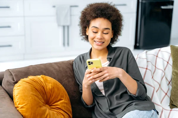Using gadgets. Lovely positive trendy african american young woman, sits on a sofa against the background of the kitchen, using smart phone, chatting in social media, messaging, shopping online, smile