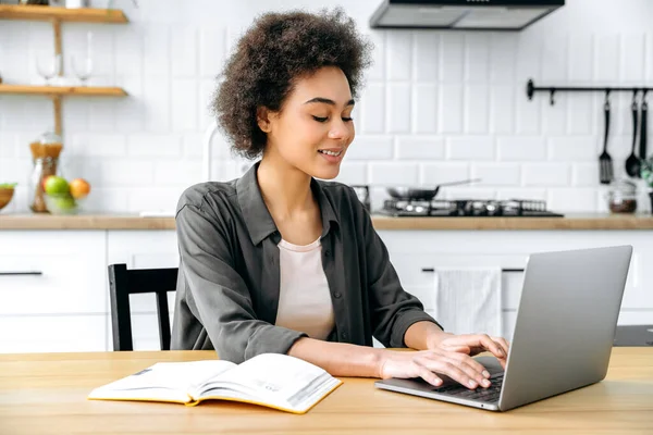 stock image Working on a project online. Successful positive african american girl with short curly hair, using a laptop in a kitchen, typing on a keyboard, studying, working, develops a plan and strategy, smile