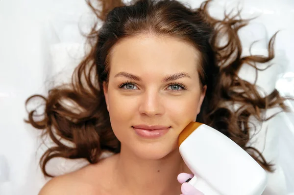 Close-up of a positive lovely caucasian young woman lying in a beauty salon during the procedure for removing unwanted hair using laser hair removal. Laser hair removal on the face. Salon procedures