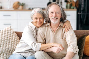 Orta yaşlı evli beyaz çift, gri saçlı eşler oturma odasında birlikte vakit geçiriyorlar, karı koca kanepede oturuyor, sarılıyorlar, kameraya bakıyorlar, gülümsüyorlar. Mutlu emeklilerin portresi.