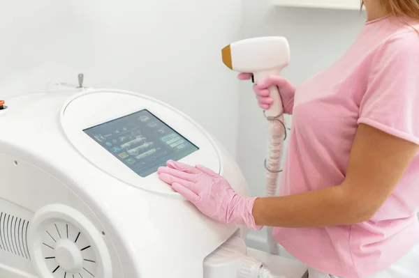 stock image Laser machine. A woman beautician in a special suit and in pink gloves stands in a special room of a beauty salon carrying a laser machine for laser hair removal, selects a settings on touch screen