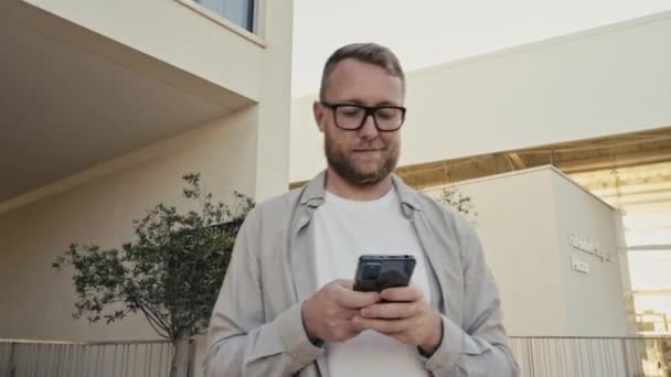 カジュアルな服装で魅力的な白人男性は 屋外に立って 彼のスマートフォンを使用して メッセージを書き込み ソーシャルメディアで友人とテキストを書き込み 情報を検索し 電子メールを入力し 電話画面を見て — ストック動画