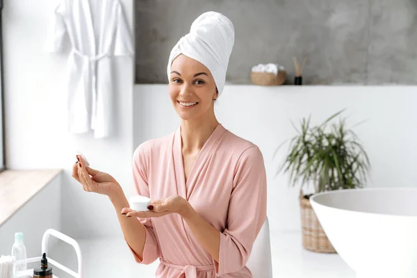 Beautiful happy caucasian brunette long haired woman in bathrobe with towel on head, sit in a bathroom, holds a white cotton pad to cleanse her sensitive skin, uses skin care products, smile at camera