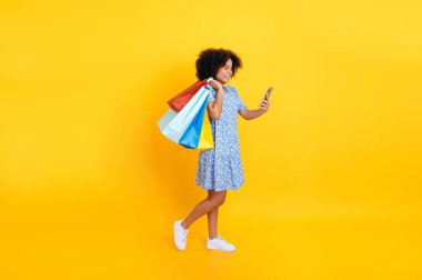 Modaya uygun, pozitif Brezilyalı ya da İspanyol kıvırcık kadın, mavi bir elbise içinde, elinde kağıt torbalarla akıllı telefon kullanıyor, ekrana bakıyor, gülümsüyor, izole edilmiş sarı arka planda duruyor.