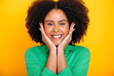 Yakından çekilmiş güzel, Afro-Amerikan ya da Brezilyalı kıvırcık saçlı, kahverengi gözlü, yeşil gömlekli, kameraya bakan, güler yüzlü, dost canlısı, izole turuncu arka planı olan genç bir kadının fotoğrafı.