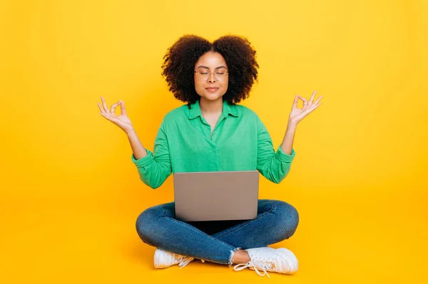 stock image Full-length photo of calm relaxed african american or brazilian curly woman, stylishly dressed, sits on a yellow background with a laptop, meditate with eyes closed, take care of mental health, smiles