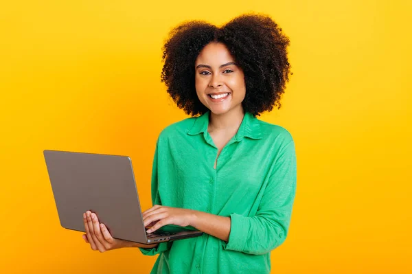 stock image Positive beautiful brazilian or african american curly haired woman, dressed in a green shirt, holding an open laptop in hand, looking at camera, smiling, standing over isolated orange background