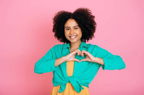 stock image Beautiful friendly kind sincere brazilian or latino curly woman showing heart gesture with hands near chest, demonstrating love, peace, support, smiling at camera, stand on isolated pink background