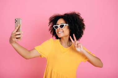 Mutlu güzel, modaya uyan Brezilyalı ya da İspanyol kıvırcık kadın, blog yazarı, güneş gözlüğü takan ve elbise giyen, akıllı telefonunda özçekim yapan, poz veren, gülümseyen, barış işareti gösteren, soyutlanmış pembe arka plan