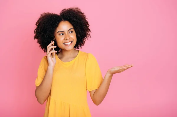 stock image Glad lovely hispanic or brazilian woman, in yellow sundress, talking on mobile phone with friend or client , looks away, points with hand aside at empty space, smile, stand on isolated pink background