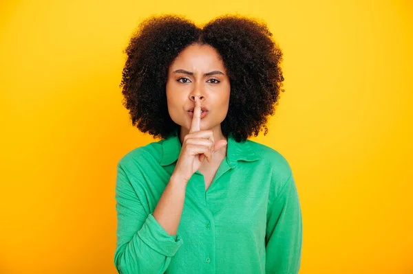 Secret, mystery. Photo of pretty brazilian or latino woman, wearing casual wear, showing shh sign with forefinger touching her lips, stop talking, looks at camera, stand on isolated yellow background