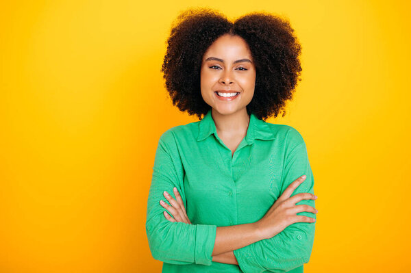 Portrait of a positive gorgeous hispanic or brazilian curly haired woman, wearing a green casual shirt, looking at the camera, smiling, standing with arms crossed on an isolated yellow background