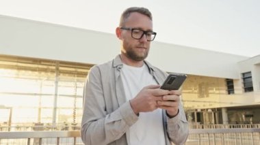 Pozitif beyaz adam dışarıda duruyor, akıllı telefonunu kullanıyor, mesaj yazıyor, arkadaşlarıyla mesajlaşıyor, internette haber okuyor, bilgi arıyor, telefon ekranına bakıyor, gülümsüyor.