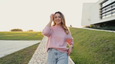 Güzel, modaya uygun, beyaz, sarışın, şık giyinmiş, gün batımında dışarıda yürüyen, kameranın önünde mutlu bir şekilde poz veren güzel bir kadının videosu.