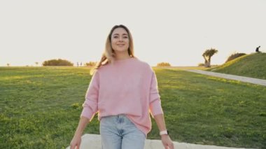 Parkta yürüyen bir kadın. Muhteşem, modaya uygun, beyaz, sarışın, şık giyinmiş, gün batımında dışarıda yürüyen, kameranın önünde mutlu bir şekilde poz veren,