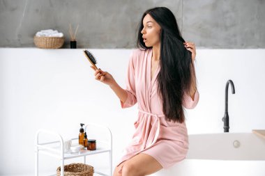 Kafası karışmış, uzun siyah saçlı beyaz bir kadın, banyoda oturuyor, elinde bir tarak tutuyor, saçını taradıktan sonra şaşkınlıkla ona bakıyor.