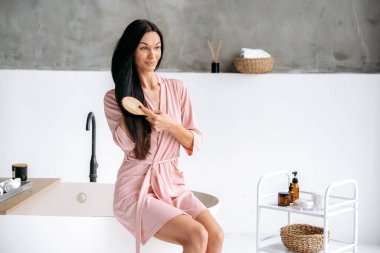 Uzun saçlarla savaşıyor. Güzellik rutini konsepti. Beyaz tenli, uzun saçlı, bornoz giyen, banyoda oturan, kalın siyah saçlarını ahşap tarakla tarayan, gülümseyen bir kadın.