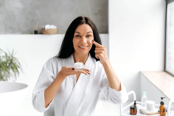Daily skin moisturizing treatments. Beauty concept. Beautiful caucasian brunette woman, in a white bathrobe, holding a jar of cream in hand, applies a moisturizing anti-aging cream on her face, smiles