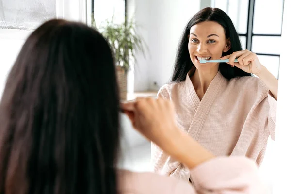 Dental care, morning routine. Lovely caucasian woman in a bathrobe, brushing her teeth after sleeping standing in front of a mirror in the bathroom, taking care of oral hygiene to avoid caries