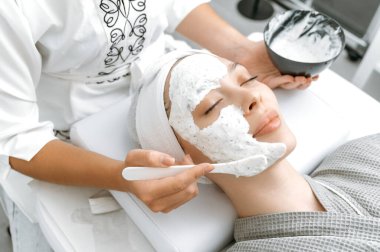 Güzellik uzmanı, güzellik salonunda gözleri kapalı yatan güzel bir kadının yüzüne nemlendirici bir yaşlanma önleyici maske takıyor. Cilt bakımı, güzellik prosedürleri, spa günü.